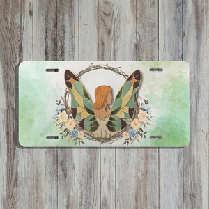 May include: License plate featuring a fairy design. The artwork depicts a woman with red hair and butterfly wings, surrounded by a wreath of branches and flowers. The background has a watercolor effect with shades of green and white.
