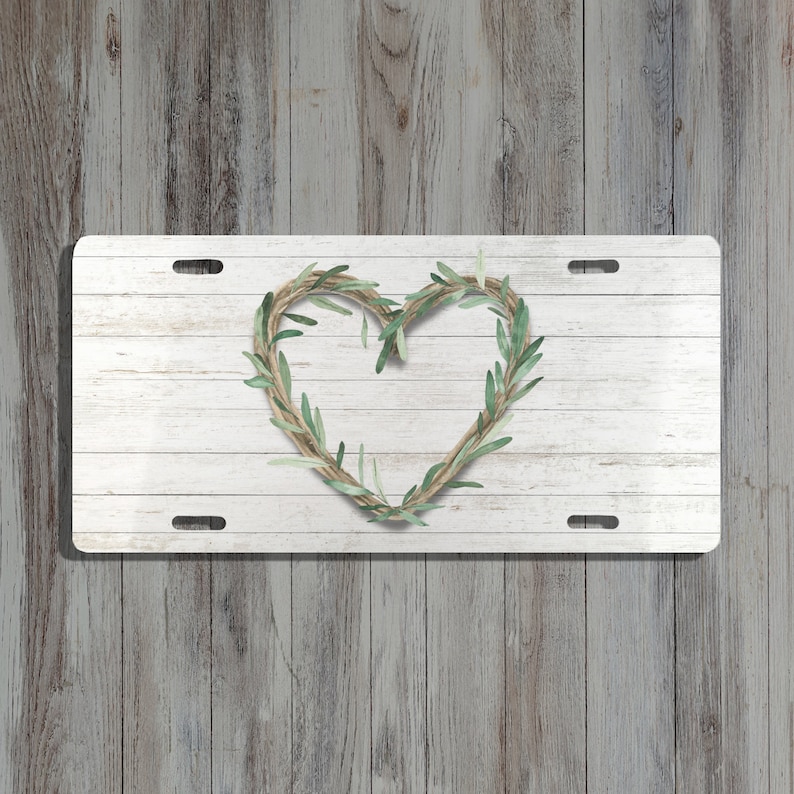 May include: Rectangular license plate with a heart-shaped wreath of twine and green leaves. The design is set against a white, distressed wood-effect background. Includes four mounting holes.