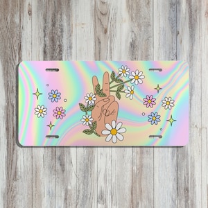May include: License plate with a holographic background featuring a hand making a peace sign with daisies and leaves. Additional daisies, stars, and flowers are scattered around the hand.