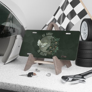 Pode incluir: Uma placa de matr&iacute;cula verde com uma borboleta branca e o texto "As Above So Below" em uma fonte vintage. A borboleta voa ao redor de uma lua cheia com flores.
