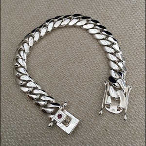 Może przedstawiać: Srebrna bransoletka z łańcuszkiem typu curb i zapięciem ozdobionym małym czerwonym kamieniem.