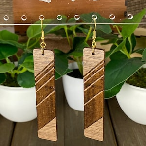 Könnte beinhalten: Ein Paar Holzohrringe mit geometrischem Muster. Die Ohrringe sind aus dunkelbraunem Holz mit einer helleren Holzeinlage gefertigt. Die Ohrringe hängen an goldenen Haken.