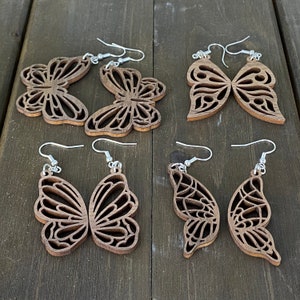 Puede incluir: Conjunto de seis pendientes de mariposa de madera con ganchos plateados. Las mariposas están talladas de forma intrincada y tienen un acabado de madera natural.