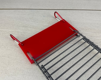 Puede incluir: Una rejilla de parrilla de metal roja con un fondo de malla de alambre y dos clips rojos en los lados.