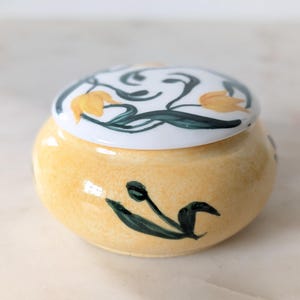May include: A round ceramic trinket box with a yellow base and a white lid. The yellow base features a hand-painted dark green tulip motif. The white lid is decorated with a pattern of yellow tulips and dark green leaves and stems.