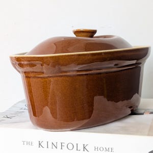 May include: A glossy, oval-shaped brown ceramic casserole dish with a matching lid. The dish features a smooth, reflective finish and a slightly flared rim. The lid has a small, centred knob handle, and the overall design is simple and functional.