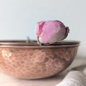 May include: A round, hammered copper bowl with a metallic, reddish-brown finish and a distinct textured surface. Resting inside the bowl is a single, soft pink peony flower bud with delicate, slightly wilted petals at the top edge.