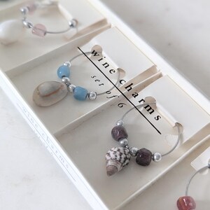 May include: A set of six wine charms in a white box. Each charm features a different design, including a seashell, a blue stone, and a pink stone. The box has the text "wine charms set of 6" printed on it.