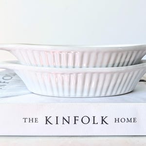 May include: Two stacked white ceramic baking dishes with fluted sides and small handles on each end. The dishes are resting on a white book with the text THE KINFOLK HOME printed in black on the front cover, featuring a clean and simple design.