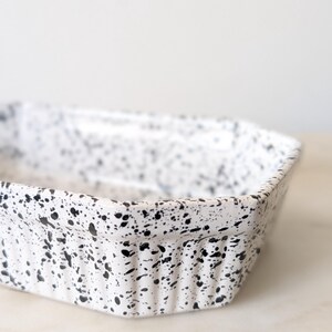 May include: A rectangular ceramic baking dish featuring a white base with a black speckled pattern. The exterior walls of the dish have a vertical ribbed texture, and the rim is slightly flared, providing a functional design for kitchen use.