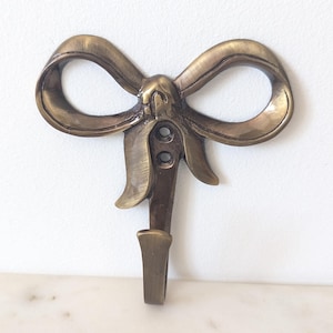 May include: A wall-mounted hook crafted from antique-toned brass, featuring a decorative bow design at the top. The hook includes two vertical screw holes centred within the bow structure, providing a functional and sturdy support for hanging items.