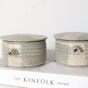 May include: Two cylindrical ceramic lidded jars with a speckled grey and off-white glaze. Each jar features a small, arched handle on the side. The jars are resting on a white book with the text THE KINFOLK HOME printed on the spine.