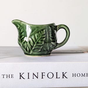 Puede incluir: Una pequeña jarra de leche de cerámica verde oscuro con un patrón de hojas de helecho en relieve en el exterior. La jarra tiene un asa curva y un pico vertedor, con un acabado brillante que refleja la luz sobre su superficie texturizada.