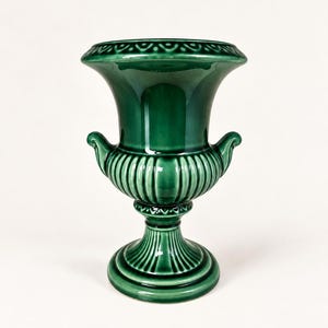 May include: A glossy emerald green ceramic urn vase featuring a wide flared rim with a decorative scalloped pattern, a ribbed bulbous body, and two small scrolled handles on the sides, all supported by a sturdy pedestal base with vertical fluting.