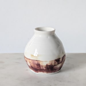 May include: A small, round ceramic vase with a glossy white glaze on the upper portion and a wide, horizontal band of dark reddish-brown, brush-stroke style pigment around the lower base, featuring a narrow opening and a smooth, curved body.