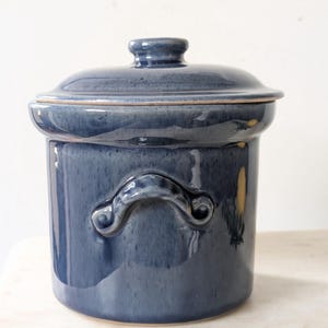 May include: A cylindrical ceramic crock with a glossy, deep blue glaze. The container features two curved side handles and a matching lid with a central knob. The surface shows subtle variations in colour depth, characteristic of a glazed finish.