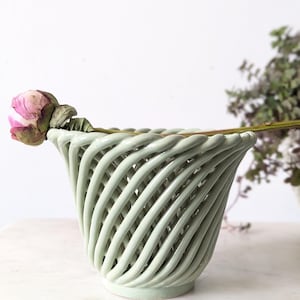 May include: A pale green ceramic vase with a woven pattern. A dried pink flower sits on the rim. The vase has a wide opening and a circular base. The background is a soft white.