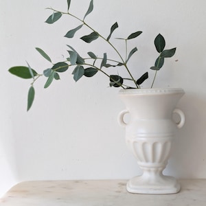 May include: A white ceramic urn-style vase with two side handles and a fluted base, containing a single stem of green eucalyptus leaves with slender, oval-shaped foliage extending outwards from the top opening of the vessel.