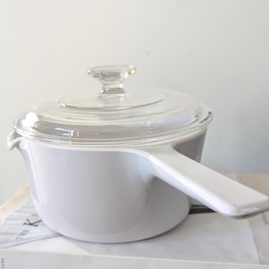 May include: White enamel saucepan with a glass lid and a white handle. The saucepan is sitting on a book titled "The Kinfolk Home".