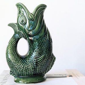 May include: A glossy dark green ceramic pitcher shaped like a fish with its mouth open. The pitcher features detailed scale textures, prominent fins, and a circular opening between the curved tail and the body, resting on a circular base.
