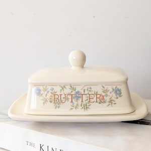 May include: A cream-colored ceramic butter dish with a rectangular base and a matching lid featuring a knob handle. The front of the lid is decorated with a floral pattern of small blue flowers and green leaves, with the word BUTTER in brown text.