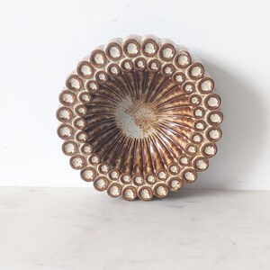 May include: A round, ceramic decorative bowl with a brown and beige color scheme. The bowl features a textured, radial design with a series of small, circular indentations around the rim. The interior has a speckled, light brown appearance.