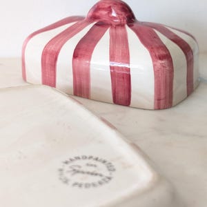 May include: A ceramic butter dish featuring a rectangular base and a lid with a rounded handle. The lid is decorated with vertical, hand-painted dark red stripes on a cream-coloured background. The base has a circular stamp reading Handpainted.
