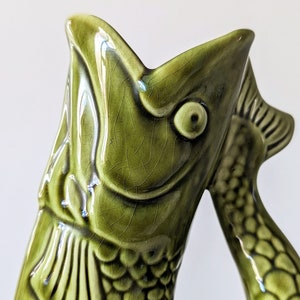 May include: A glossy olive green ceramic pitcher shaped like a fish with its mouth wide open. The surface features a fine crackle glaze finish, detailed scales, and a prominent eye. The handle is formed by the curved tail of the fish.