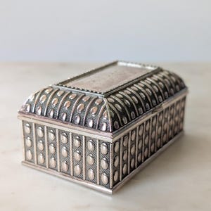 May include: A rectangular silver-toned metal box with a hinged lid. The exterior features a repeating pattern of vertical columns containing raised oval studs set against a textured, dark-toned background. The top lid has a smooth, flat center.