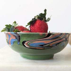 May include: A green ceramic bowl with a swirled pattern of blue, orange, and black. The bowl is filled with fresh strawberries. The bowl is on a white surface.