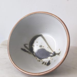 May include: A small, ceramic bowl with a white interior and a bronze-colored rim. The interior features a dark gray and black abstract design. The bowl is likely handmade and suitable for serving small portions of food or as a decorative item.