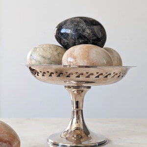 May include: A silver-toned metal pedestal bowl with a decorative cut-out rim pattern, holding a collection of smooth, polished stone eggs in shades of black, cream, and tan. An additional tan stone egg rests separately to the left of the bowl.