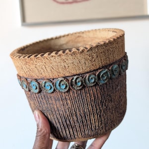 May include: A cylindrical ceramic planter with a rough, dark brown textured exterior featuring vertical grooves. The upper rim has a wide, light tan band with a notched edge, accented by a row of small, circular turquoise blue spiral motifs.