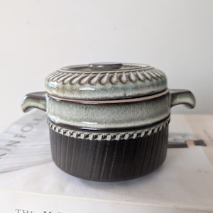 May include: A ceramic lidded pot with two side handles, featuring a two-tone glaze. The upper section and lid are a light, mottled blue-green, while the lower body is a dark brown with vertical ribbed textures and a decorative beaded border.