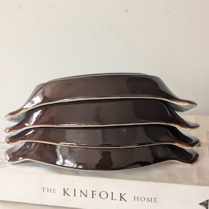 May include: A stack of four dark brown ceramic bowls with small side handles. Each bowl features a glossy finish and a light-coloured rim. The bowls are stacked vertically, showcasing their curved shape and smooth, reflective surface texture.