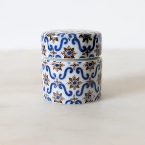 Pode incluir: Um pequeno recipiente cilíndrico de cerâmica com tampa, apresentando um padrão repetitivo de motivos florais em forma de estrela castanha e volutas em forma de S azuis sobre um fundo branco. O recipiente tem um acabamento liso e brilhante.