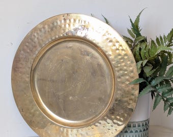 May include: A round, gold-colored metal tray featuring a hammered texture along its wide rim and a smooth, flat center. The surface of the tray displays a reflective, metallic finish with visible signs of wear and light scratches across the metal.