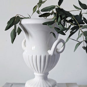 May include: A white ceramic urn-style vase with a fluted body, two side handles, and a square base. The vase features a subtle crackle glaze finish and contains a sprig of green eucalyptus leaves, resting atop a book titled The Kinfolk Home.