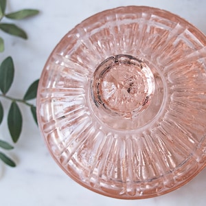 May include: A round, pale pink glass lid featuring a radial ribbed pattern and a central circular knob handle. The glass is translucent with a smooth, reflective surface, showcasing a symmetrical design that radiates from the center to the edge.