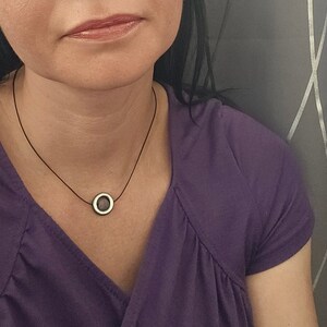 Könnte beinhalten: Eine einfache Halskette mit einem runden Silberanhänger an einem schwarzen Band. Der Anhänger hat eine dunkelgraue Mitte.