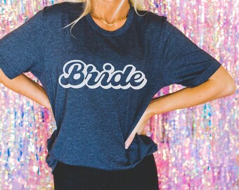 May include: A navy blue t-shirt with the word "Bride" in white, cursive lettering. The shirt is a crew neck style and has short sleeves. The background is a shiny, pink and silver foil.