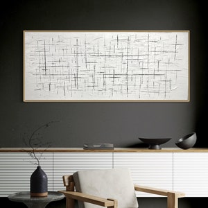 Puede incluir: Una pintura de lienzo blanco con líneas negras que crean un patrón de cuadrícula cuelga sobre un gabinete blanco con una parte superior de madera. Una mesa auxiliar de piedra gris con un jarrón de ramas secas se encuentra junto a un sillón gris claro con patas de madera.