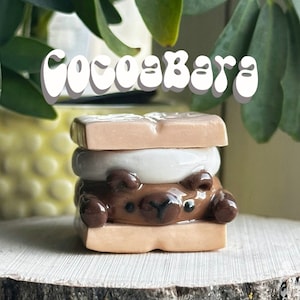 May include: A brown and white ceramic s'more with a bear face. The s'more is made of two graham cracker squares, a white marshmallow, and a chocolate bar.