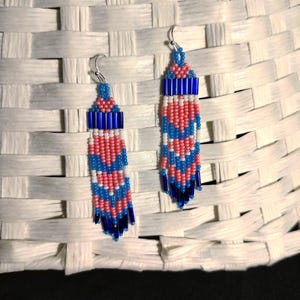 May include: A pair of dangle earrings with a blue, pink, and white beaded design. The earrings are made with small seed beads and have a silver hook.