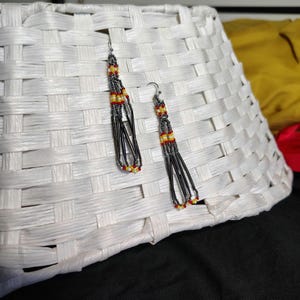May include: A pair of long, beaded earrings with black, red, and yellow beads. The earrings are hanging from a white wicker basket.