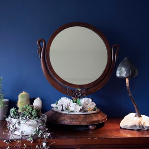 May include: A vintage wooden vanity mirror with swan-shaped handles and a white floral design on the base. The mirror is on a wooden table with a potted succulent plant and a mushroom-shaped lamp.