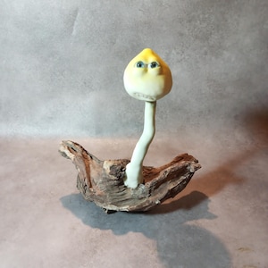 Pode incluir: Uma figura esculpida que se assemelha a um cogumelo com um chapéu amarelo pálido e um caule branco longo e fino. O chapéu apresenta dois olhos azuis e uma boca pequena. A figura está fixada num pedaço de madeira flutuante castanha texturada.