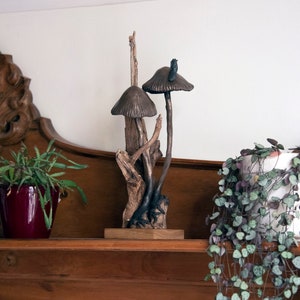 May include: A wooden sculpture of two mushrooms with brown caps and stems, set on a wooden base. The sculpture is on a wooden shelf with a green plant in a red pot and a trailing green plant in a white pot.