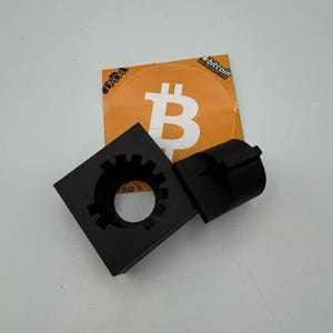 May include: Two black plastic 3D printed parts, one square with a gear-shaped hole and the other a cylinder with a slot. The parts are on a white surface with a sticker of a bitcoin logo on a yellow background.
