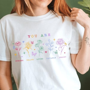 May include: White t-shirt with a colorful floral design and the text "YOU ARE" above the flowers. The flowers are labeled with the words "strong", "brave", "loved", "capable", and "smart".